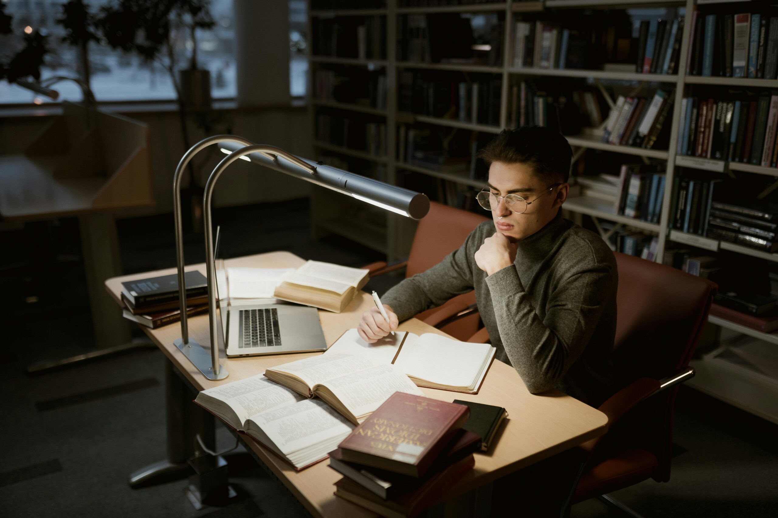 A young adult studying at a library desk with books and laptop under warm lighting.